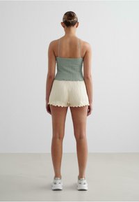Top scurt verde cu dungi, cu bretele subțiri, asortat cu pantaloni scurți crema plisati cu margini zimțate. Modelul poartă adidași albi.