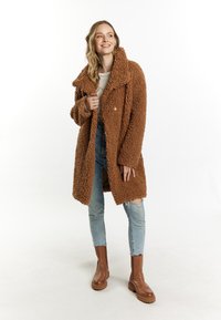 Brun teddy frakke med en stor krave, tekstureret stof og trykknapper. Båret over en cremefarvet top og parret med slidte jeans og brune støvler.