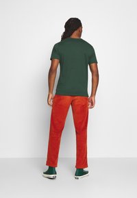 Tmavě zelené tričko s krátkým rukávem v kombinaci s oranžovými manšestrovými kalhotami; model má na sobě zelené vysoké tenisky. Pozadí je jednoduché bílé.