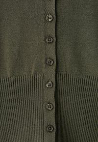 Gros plan d’un tissu tricoté vert olive avec six boutons foncés alignés verticalement et une texture côtelée en bas.