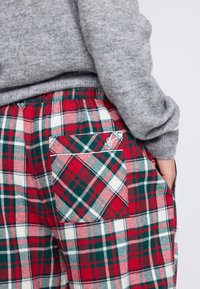 Persoon in rode, groene en witte geruite broek met een achterzak en een lichtgrijze gebreide trui, met een hand in de zijzak gestoken.