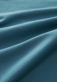 Tealfärgad tyg med en slät textur, med mjuka, flödande draperingar och ett enhetligt mönster på ytan som framhäver dess elasticitet.