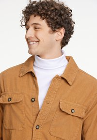 Bruine ribfluwelen shirt met een button-down kraag, twee borstzakken en zwarte knopen, gelaagd over een witte coltrui. Geweven stof.