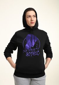 Persona con un hoodie nero e una grafica viola di una ragazza con il testo "Sub-Urban Gothic", mani in tasca, guarda in alto a sinistra.
