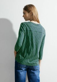 Zelený top s vzorem a třičtvrtečními rukávy, se geometrickým designem v různých odstínech zelené a volným střihem. Nosený s modrými džínami.