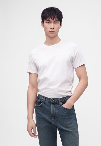 Jeune homme aux cheveux foncés portant un t-shirt blanc uni et un jean bleu, debout avec une main dans la poche sur un fond blanc.