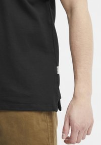 Černé bavlněné tričko s krátkými rukávy, postranními rozparky a malou cedulkou na levé straně. Hladká struktura, volný střih a jednoduchý design.