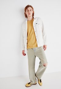 Chaqueta de mezclilla blanca sobre una camiseta mostaza, combinada con jeans verdes claros desgastados y zapatillas amarillas con rayas negras. Diseño simple y casual.