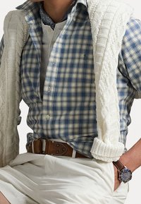 Sininen ja valkoinen ruudullinen paita, jonka päällä on valkoinen neulepusero hartioilla, ruskea nahkavyö ja tumma kello ranteessa.