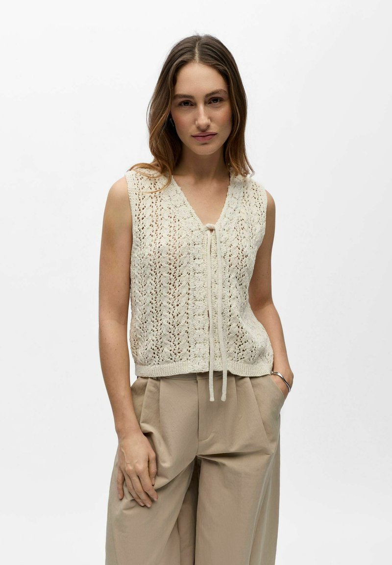 Femme aux cheveux longs portant un gilet en maille crème sans manches noué à l'avant et un pantalon beige taille haute plissé, debout avec une main dans la poche.