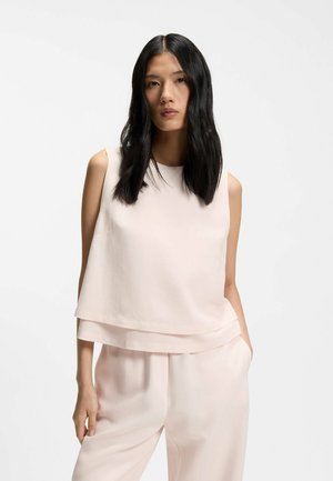 Pitkähiuksinen nainen, jolla on mustat hiukset, pukeutuneena vaaleanpunaiseen hihattomaan toppiin ja siihen sopiviin väljihin housuihin, seisoo yksivärisen vaalean taustan edessä.