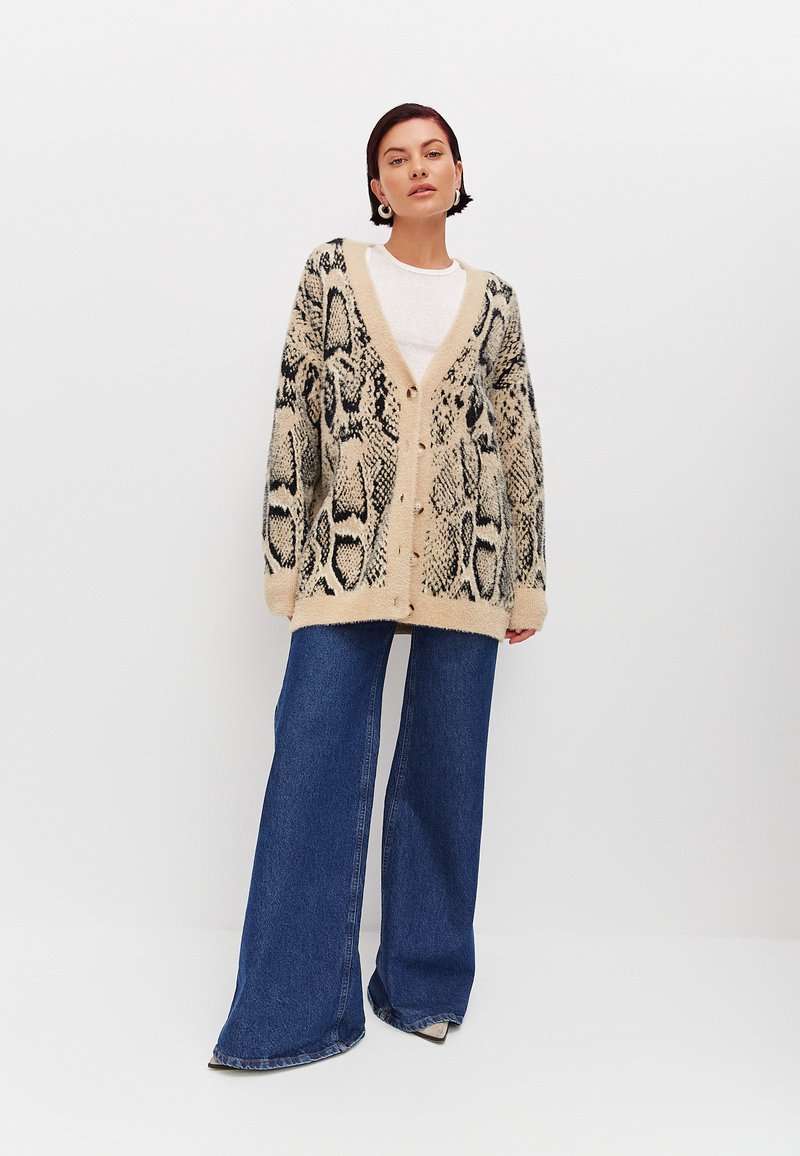 Gebreide cardigan met een beige achtergrond en een zwart slangenpatroon, met een drop shoulder en vijf knopen. Gecombineerd met wijde blauwe jeans.