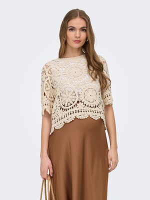 Ung kvinne iført beige heklet topp med sirkulære mønstre og brun skjørt, holder flettede tauhåndtak mot en ensfarget bakgrunn.