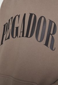 Felpa marrone con una stampa di testo nero prominentemente curva che recita "PEGADOR." Realizzata in tessuto morbido con una texture liscia.