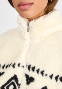 Crème-kleurige fleece jack met een hoge kraag, ritssluiting en zwarte geometrische patronen op de voorkant. Zachte textuur.