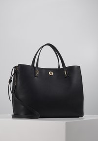 Sac à main en cuir noir avec deux poignées supérieures, une bandoulière amovible, un accent en logo circulaire doré et une finition texturée.