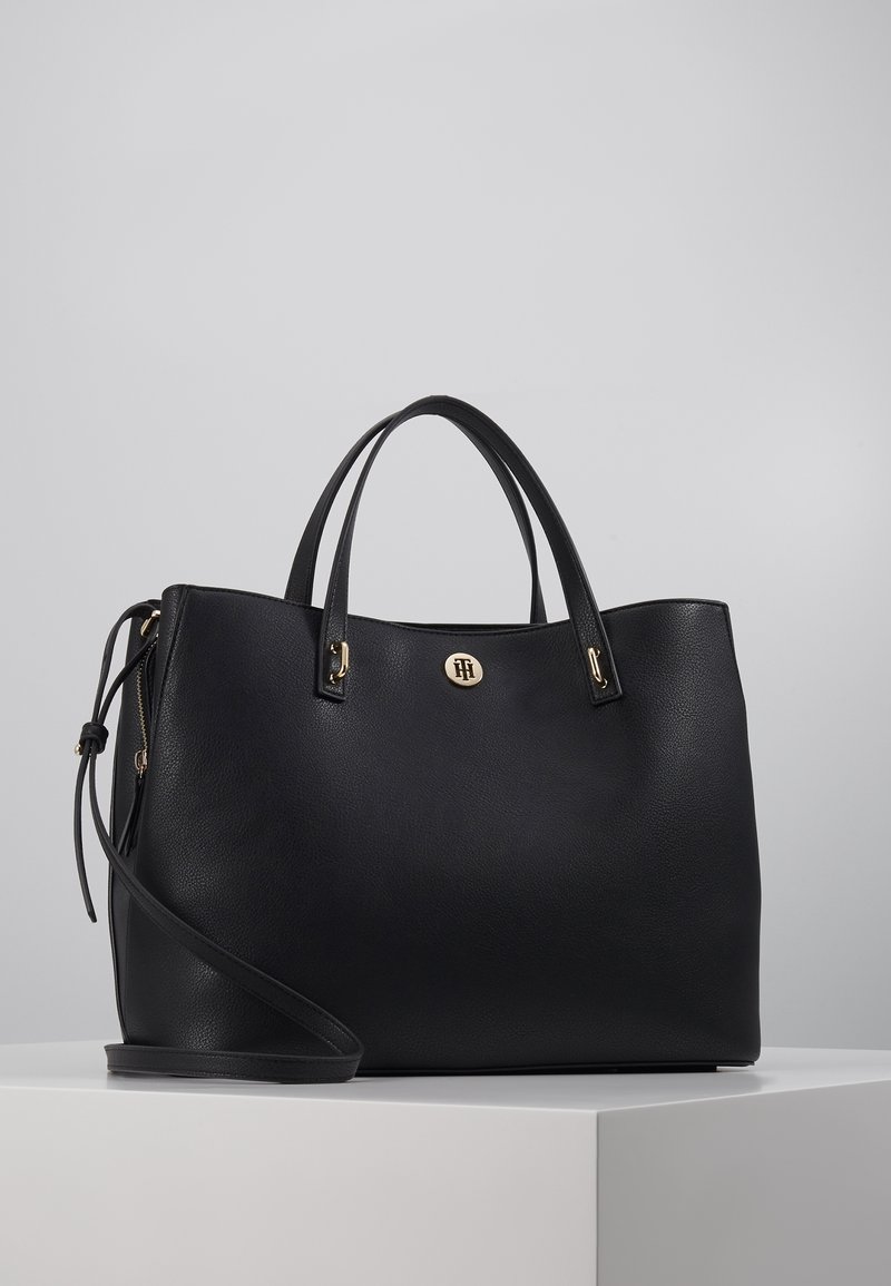 Sac à main en cuir noir avec deux poignées supérieures, une bandoulière amovible, un accent en logo circulaire doré et une finition texturée.