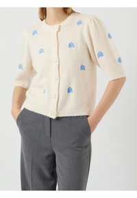 Crème cardigan met korte pofmouwen en blauwe bloemenborduursels, voorzien van een geribbelde kraag en knoopsluiting, gecombineerd met grijze broeken.