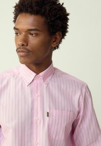 Cămașă roz deschis cu nasturi, cu dungi verticale subțiri, guler ascuțit și un buzunar pe pieptul stâng. Fabricată dintr-un material neted.