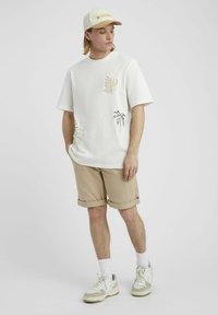Chemise blanche à manches courtes avec broderie de palmiers beiges, associée à un short beige roulé et des baskets blanches, complétée par une casquette légère.