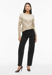 Žena stojí v béžovo-krémovom pruhovanom svetri, čiernych nohaviciach s vysokým pásom, rovného strihu, hnedých lodičkách a perlových náušniciach.