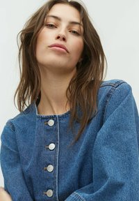 Denim jacket met een ronde halslijn, zilveren knopen en afhangende schouders. De stof is middelblauw met een gladde textuur en zichtbare naden.