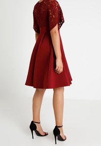 Burgundy klänning med spetsliv, utställd kjol och sömdetaljer. Bärs med svarta högklackade sandaler med ankelremmar.