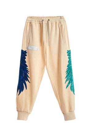 Mujer de pie con una camiseta azul suelta, pantalones jogger crema con acentos azules y verdes, y zapatos beige tipo slip-on texturizados sobre un fondo blanco.