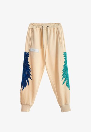 Mujer de pie con una camiseta azul suelta, pantalones jogger crema con acentos azules y verdes, y zapatos beige tipo slip-on texturizados sobre un fondo blanco.