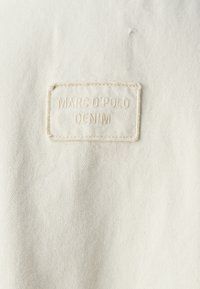 Bílá látková štítek s vyraženým textem "MARC O'POLO" a "DENIM". Štítek má prošité okraje a hladkou strukturu, umístěný na světle barevné látce.