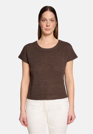 Femme aux cheveux bruns raides portant un haut en maille marron à manches courtes et un pantalon blanc, face à l'avant sur un fond uni.