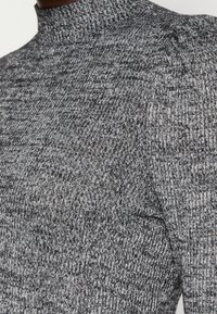 Pull côtelé gris avec un col montant et des manches bouffantes, présentant un motif en maille texturée et des accents gris foncé.