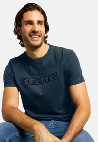 Dunkelblaues T-Shirt aus weichem Stoff, mit einem schwarzen "ELBSAND"-Logo mittig auf der Brust. Kurzarm und lässige Passform, getragen zu Jeans.