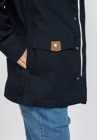 Navy blue jacket with a zip closure, featuring a patch pocket with a tan accent and silver button detail. Smooth, water-resistant fabric.