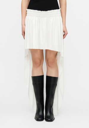 Žena oblečená v bielej sukni s vysokým a nízkym lemom, s riaseným elastickým pásom a čiernymi koženými čižmami po kolená, stojaca pred jednoduchým pozadím.