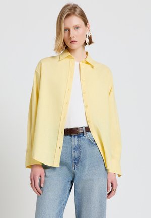 Giovane donna che indossa una camicia oversize giallo pastello con bottoni, un top bianco a costine, jeans azzurri a vita alta e orecchini vistosi.