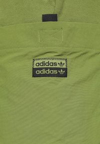 Zelená tkanina s velkou černou nášivkou loga Adidas a dvěma štítky, jeden nad druhým, obšitými cik-cak stehem.