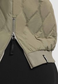 Veste matelassée beige avec une fermeture éclair argentée partiellement ouverte, montrant un tissu noir dessous et un poignet élastique côtelé à l'ourlet.