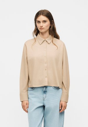 Femme aux longs cheveux bruns porte une chemise beige à manches longues boutonnée et un jean bleu clair, debout devant un fond blanc uni.