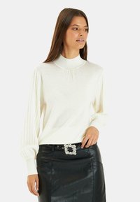 Witte gebreide trui met een hoge hals en geplooide lange mouwen. Gestyled met een zwarte leren rok met een decoratieve riemgesp.