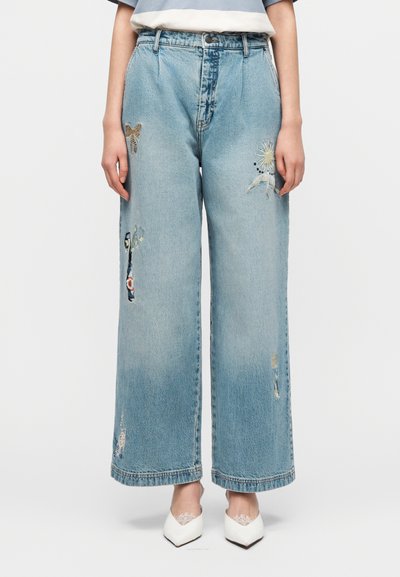 Ανοιχτό μπλε τζιν παντελόνι με φαρδιά μπατζάκια, διακοσμημένο με κεντήματα λουλουδιών και φθαρμένες λεπτομέρειες. Διαθέτει ψηλή μέση και ορατές ραφές.