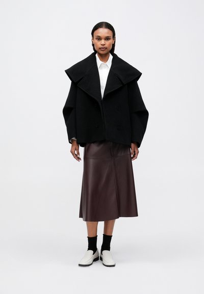 Žena nosi crni oversized kaput, bijelu majicu, tamnosmeđu midi kožnu suknju, crne čarape i bijele slip-on cipele na bijeloj pozadini.