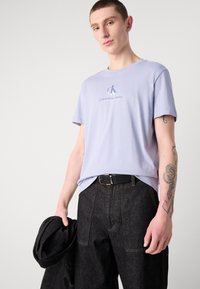 Camiseta de algodón lavanda clara con escote redondo y sutil logo de Calvin Klein en el pecho. El modelo lleva pantalones de mezclilla negros y sostiene una chaqueta negra.