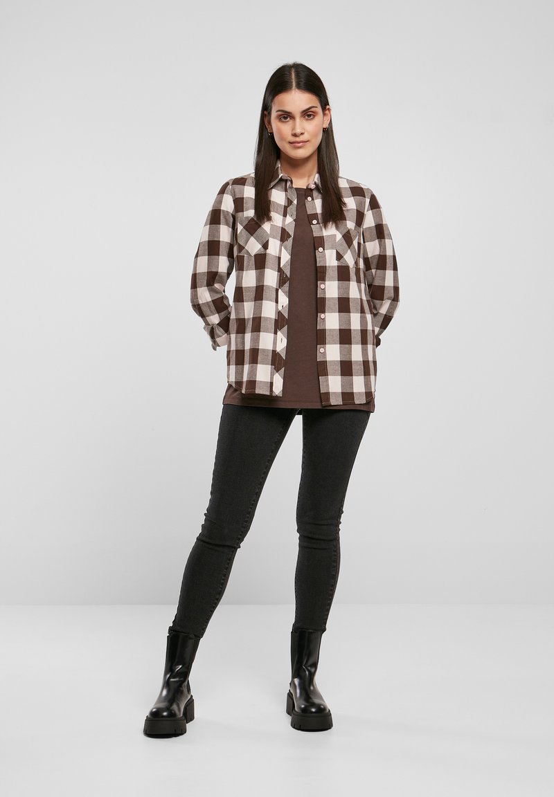 Camicia a quadri marrone e bianca con chiusura a bottoni, due tasche sul petto, indossata sopra una maglietta marrone. Abbinata a jeans skinny neri e stivali neri chunky.