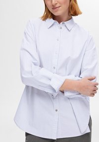 Femme portant une chemise blanche à manches longues avec de fines rayures verticales bleues, bras croisés, sur un fond clair uni.