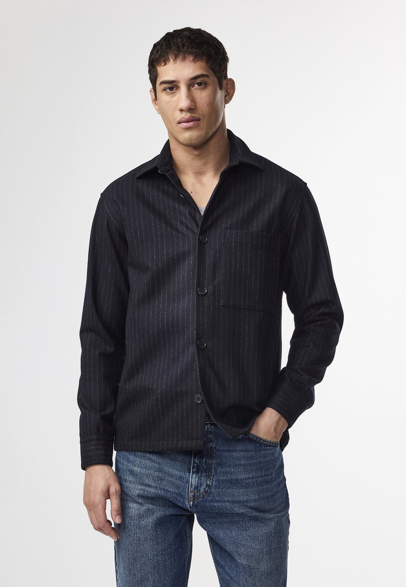 En ung man står självsäkert, klädd i en svart randig skjorta och jeans, med en neutral bakgrund som framhäver hans klädsel.