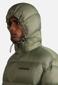Giacca verde imbottita con colletto alto e cappuccio regolabile. Presenta dettagli a pannello contrastanti e un logo nero sul petto.