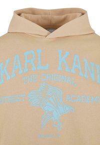Felpa beige con cappuccio regolabile. Caratterizzata da testo ricamato azzurro chiaro: "KARL KANI" e una grafica di un'aquila. Texture morbida, vestibilità casual.
