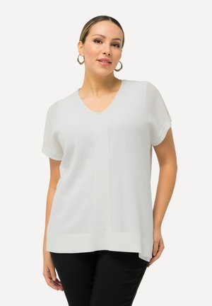 Lichtgrijze gebreide top met V-hals, korte mouwen en een textuurpatroon. Heeft een iets langere achterkant voor extra stijl. Gecombineerd met zwarte broek.
