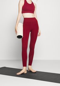 Donna in un abbigliamento sportivo rosso, che tiene un rullo di schiuma bianco con texture, in piedi a piedi nudi su un tappetino yoga nero contro uno sfondo semplice.